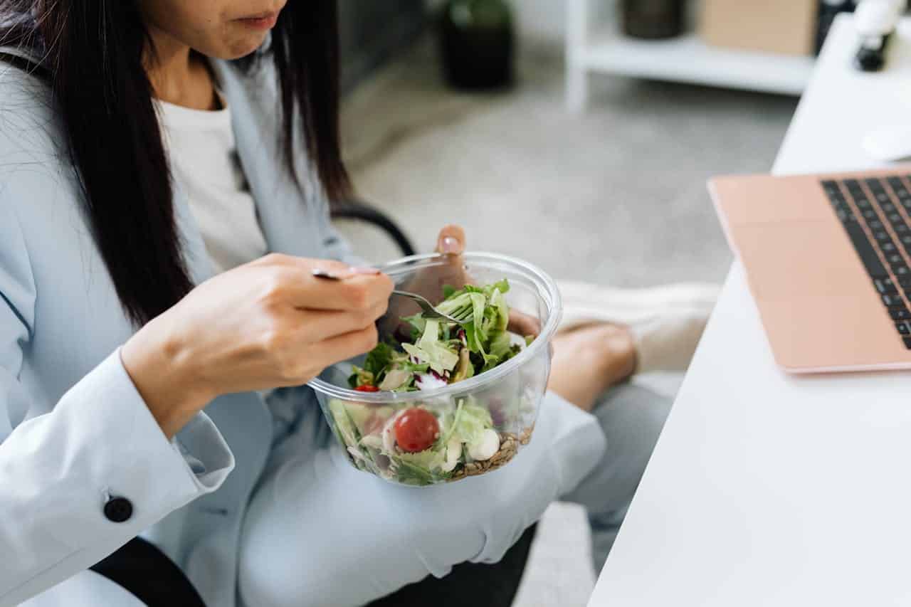 Why You Feel Sleepy After Lunch at Work and How to Stop It Woman eats a healthy lunch at work - importance of eating the right food to prevent food coma.